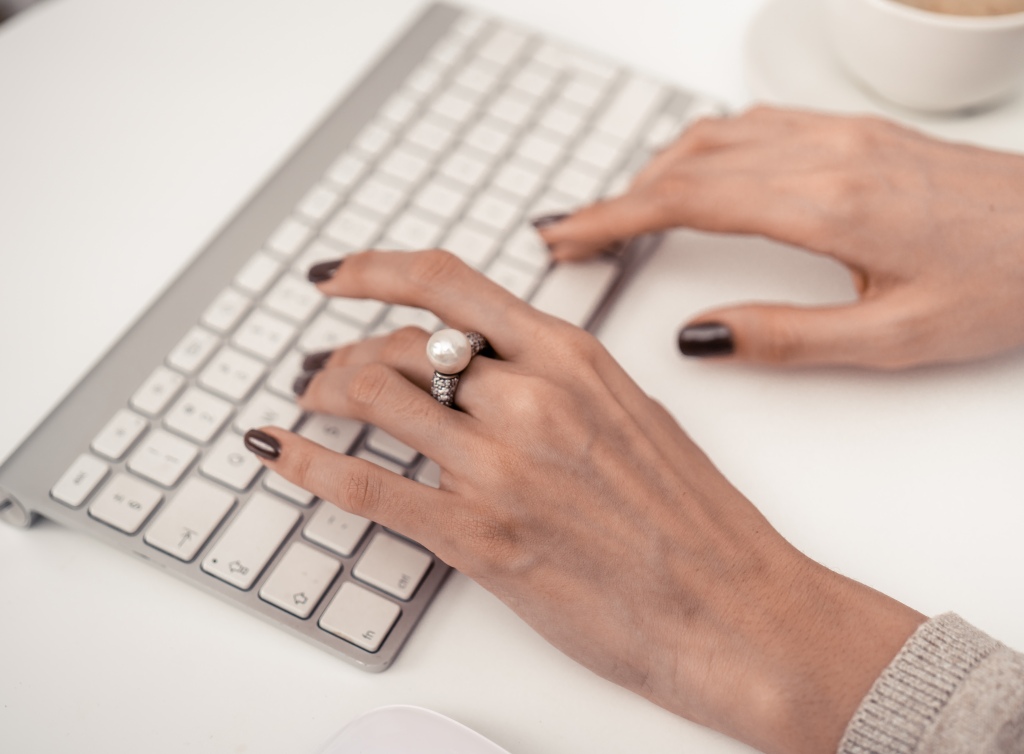Woman's manicured hands typing on wireless keyboard wearing a large pearl ring on her middle finger, a cup of coffee next to her keyboard. 